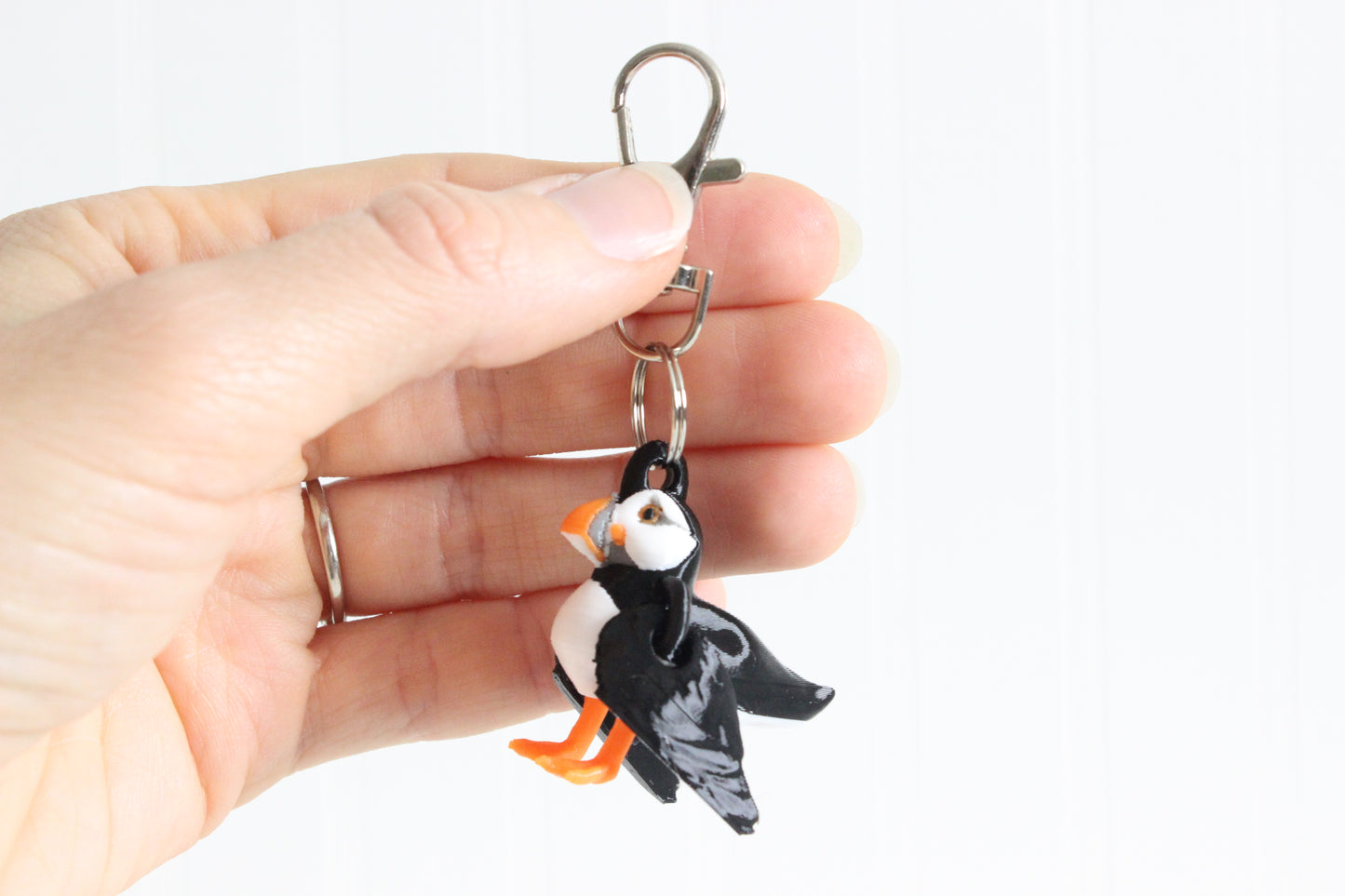Hand holding 3D-printed black, white, and orange puffin keychain to show its size, colors, and silver swivel clip.