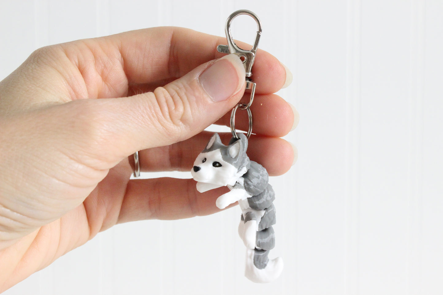 Hand holding the 3D-printed gray-and-white husky keychain to show its size, colors, and silver swivel clip.